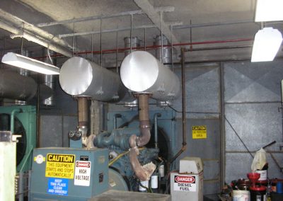 Existing pump and ceiling mounted mixing tanks beside a steel plenum wall and other miscellaneous pieces of equipment and supplies in the mechanical room