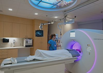 Technician in scrubs operating the large MRI machine while the patient covered with a blanket lays on the retracting table with his head inside the cylindrical machine while he receives the scan