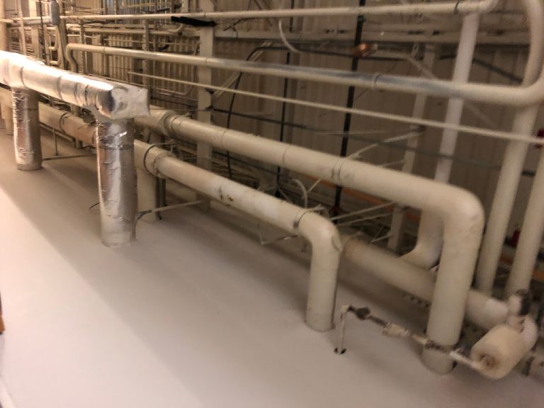 Round ductwork and large diameter chilled and hot water piping running along the mechanical room wall and turning down to penetrate the floor