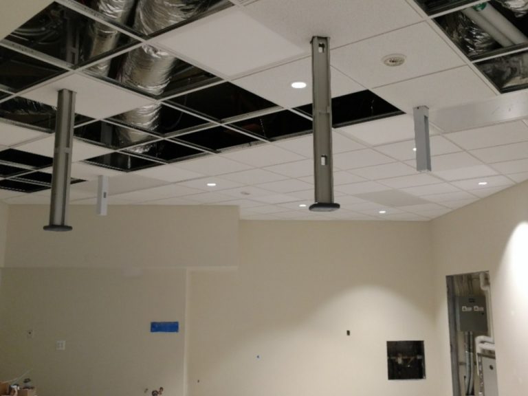Construction photo showing sections of square drop ceiling panels removed revealing circular flex duct run in the space above and several metal supports for the radiosurgery machine extending down into the exam room