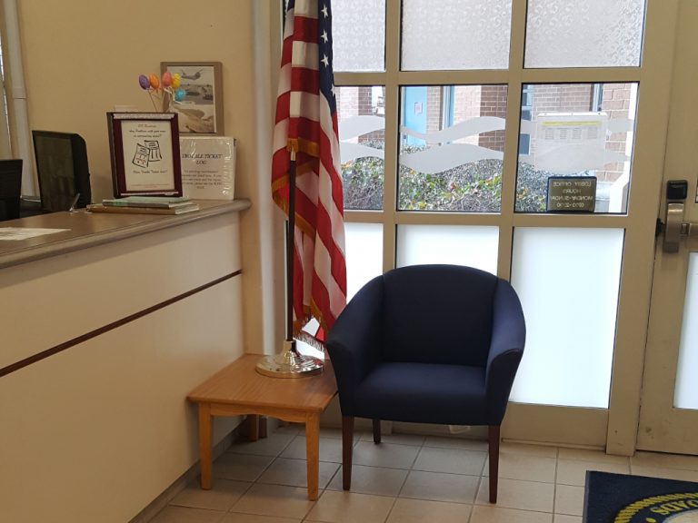 A dark blue rollback chair sitting in between the front entry door and an American flag both sitting near the front desk of the B E Q