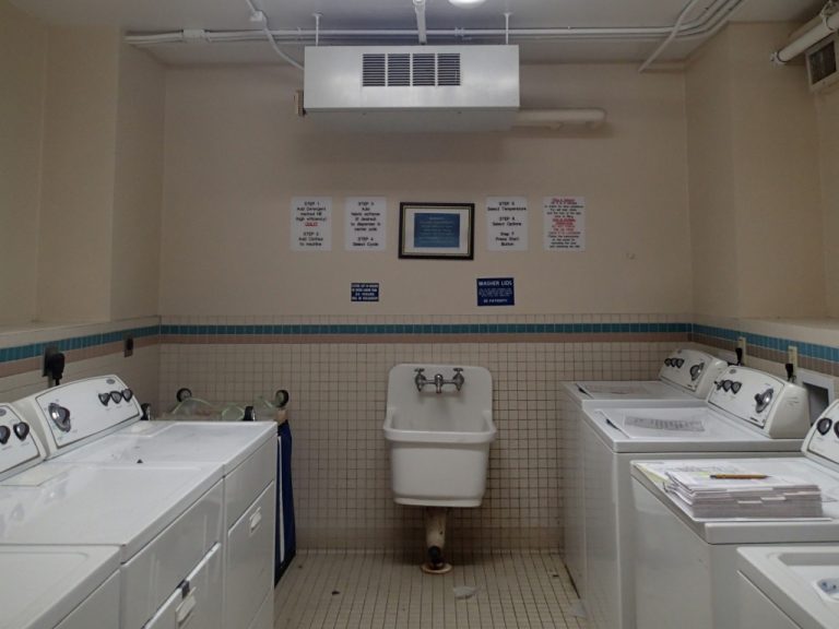 White washers and driers line both sides of the tiled laundry room and a standard white mop sink sits alone on the back wall underneath a ceiling mounted dehumidifier