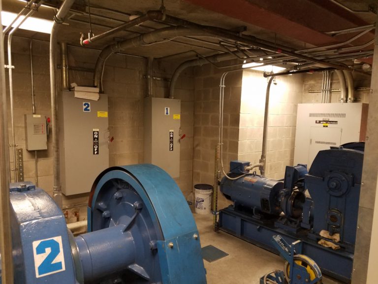 Two large floor mounted pumps and the electrical cabinets that control them mounted on the wall beyond in the mechanical room of the building