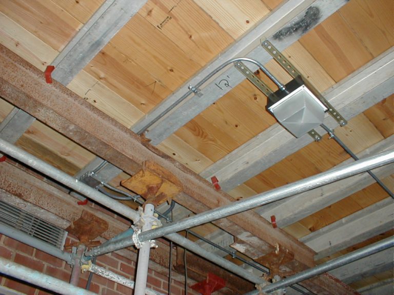 Looking up in the basement area at an emergency light and it's conduit mounted to floor beams that are being supported by a screwjack and scaffolding