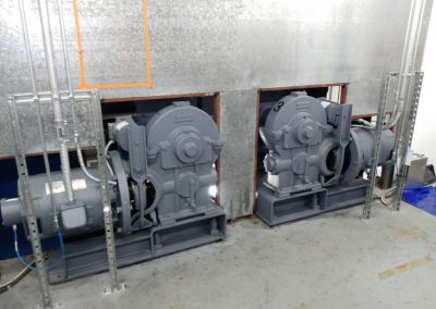 Two large pumps for the elevator system mounted to the mechanical room floor underneath a metal plenum type enclosure