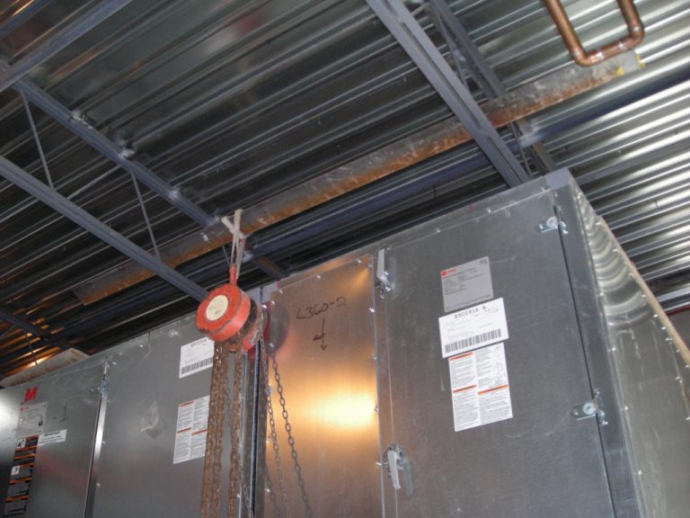 Corrugated metal roofing and metal trusses above a ceiling mounted piece of HVAC equipment and a red chain hoist