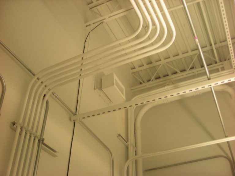 Five white conduits snaking uniformly up the mechanical room wall and across the ceiling space