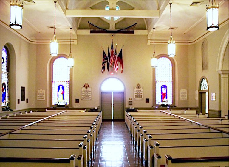 View from the front of the sanctuary of rows of pews under classical pendant lights alongside arched columns and stained glass windows