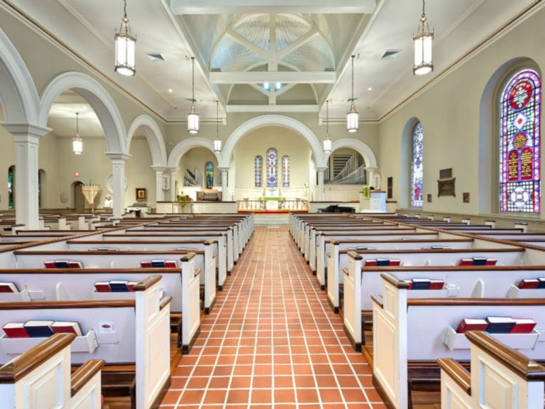 View from the rear of the sanctuary of rows of pews under classical pendant lights alongside arched columns and stained glass windows