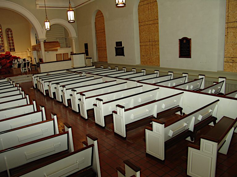 View from the rear of the sanctuary of rows of pews under classical pendant lights alongside the boarded arched windows during construction