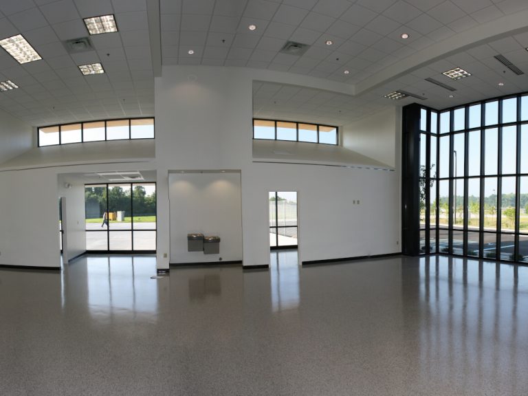 Interior view of a vaulted ceiling and a large glass facade of an empty showroom with glass accent windows and doors highlighting its polished white vinyl floor