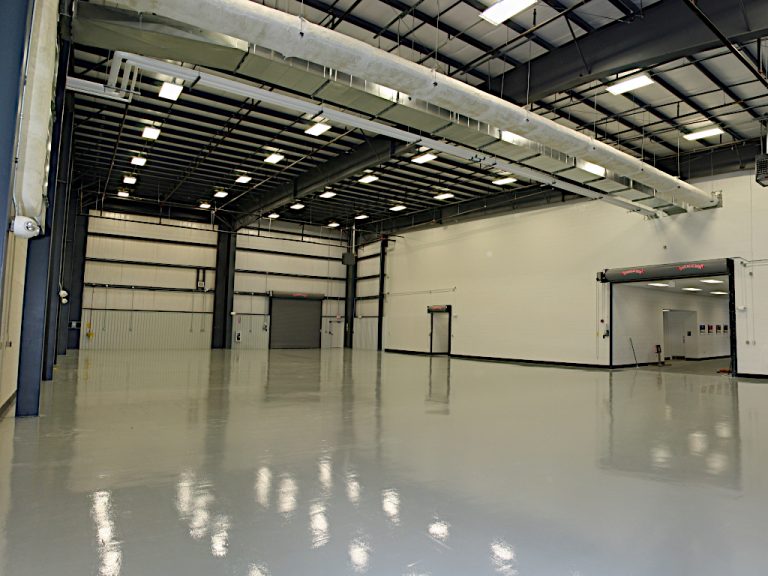 Interior view of black metal beams and ceiling mounted piping above a large empty warehouse with large access doors and a polished white vinyl floor