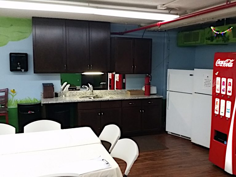 Doubletree Hotel Virginia Beach Employee Break Room with a marble counter top and dark cherry wood cabinets sitting alongside two apartment sized refrigerators, a coca-cola drink machine and two six foot break tables surrounded by several white folding chairs