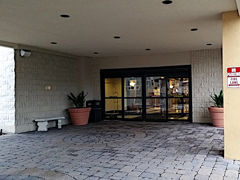 A cobblestone area underneath the portico in front of a double set of automatic sliding doors that are adorned with two large terracotta planters on either side of the entrance