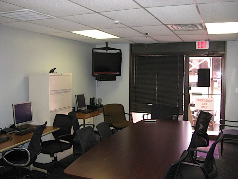 A meeting room containing a large conference table and rolling chairs sitting beside a filing cabinet and a ceiling mounted television with VCR