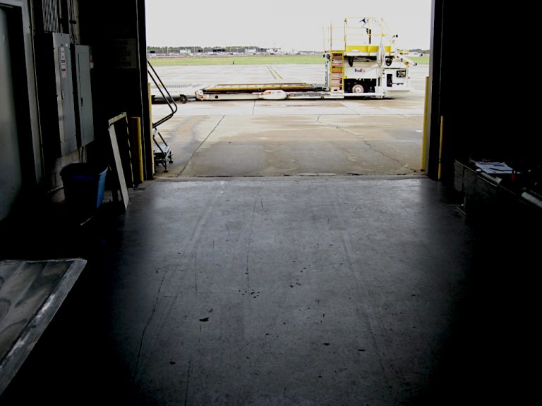 Looking out the roll up door of the warehouse into the concrete parking lot where a large FedEx load and lift type truck sits with the Norfolk airport in the distance