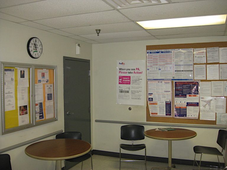 Interior of the warehouse break room with several round tables and metal and plastic chairs alongside a bulletin board and a large wall clock