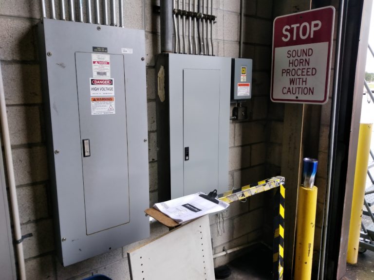 Two electrical breaker boxes mounted on the block wall beside the roll up door of the warehouse both with multiple metal conduits penetrating the top of each of them