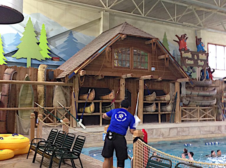 Greatwolf Lodge lifeguard standing in front of a set of large log shelves used to store rustic kayaks alongside people playing in the water of one of the swimming pools