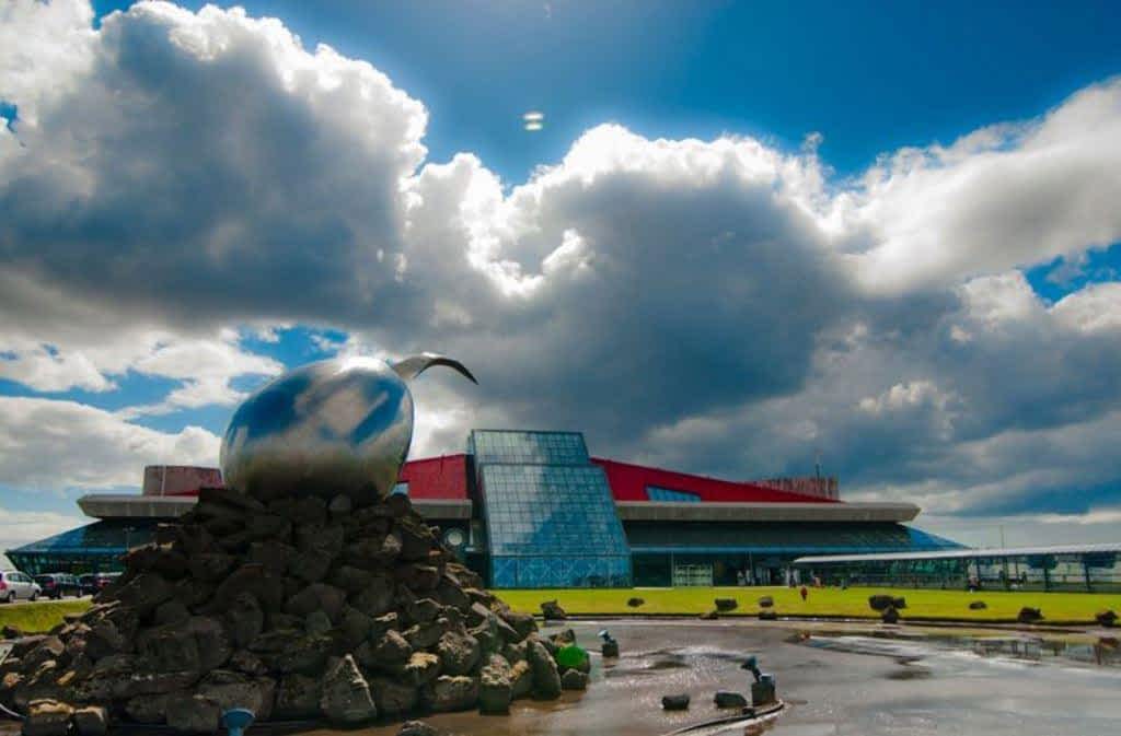Keflavik International Airport View of a giant stainless steel acorn statue perched on a pile of boulders under a blue sky with fluffy white clouds with the glass and brick Keflavik Airport in the background