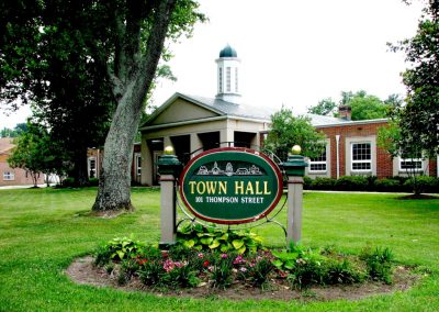 The Ashland Town Hall sign sitting in a landscaped flower bed on a freshly cut green lawn in front of the entrance to the brick town hall building