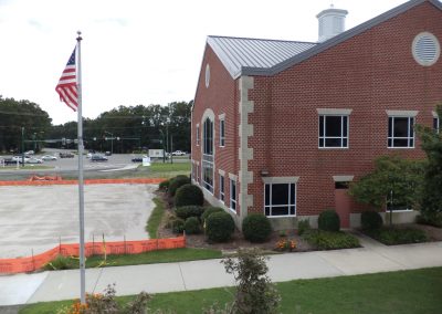 A graded dirt parking lot and landscaped walkway along side a brick and black glass building with an american flag hanging in front of it