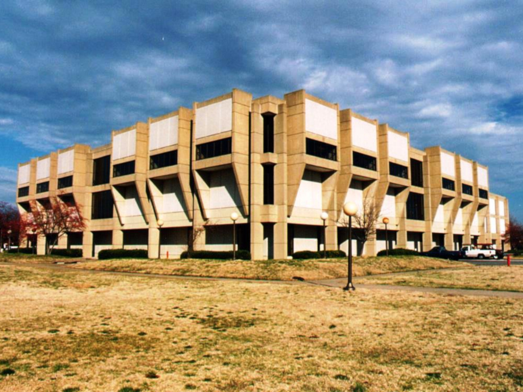 A modern concrete two story building of a higher education facility sitting in a grassy field beyond a walkway with lamp posts and a few autumn trees