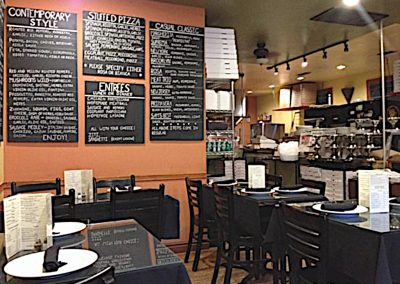 Dark wooden chairs and tables alongside several menu chalkboards mounted on the wall of a cozy little pizza restaurant