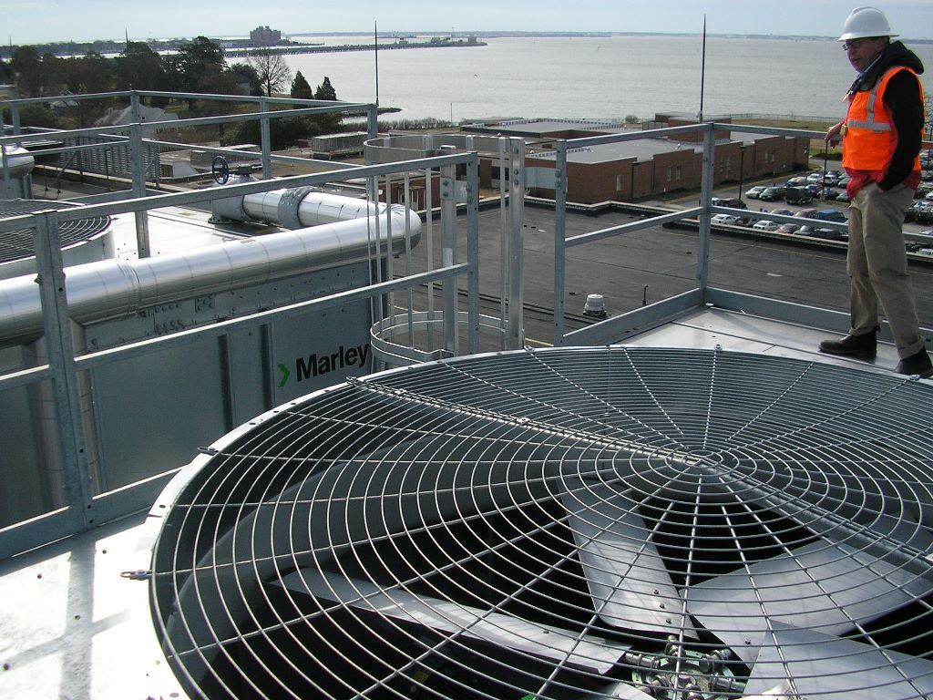 Project partner wearing a blaze orange protective vest walking along the handrail of rooftop metal platform that holds a commercial Marley brand air handling unit